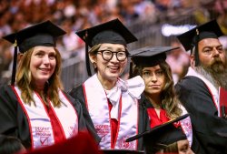 A group of Montclair State University graduates is seated at a commencement ceremony. They are wearing traditional black caps and gowns with red and white stoles that read "Degree Completion Program."