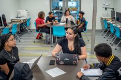 Three students gather around a small table to brainstorm, while a group of four students at another table in the background do the same.