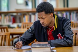 A student in a Bloomfield varsity jacket sits at a table in the library and writes notes in a notebook.