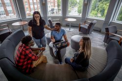 Four students gathered in a lounge talking with one another. The curved wall lined with windows behind them shows a view of downtown Bloomfield.