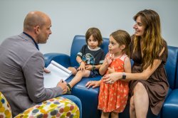 A seated man with a notepad speaks to a seated, smiling woman and a young girl as a young boy looks on.