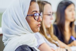A woman wearing a white hijab sits, listening, in a classroom while two other women sit, listening, in the background.