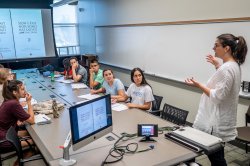 professor speaking to students gathered around a table with projection screen in the background