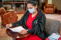A student in red scrubs and a surgical mask completes her homework in a waiting room