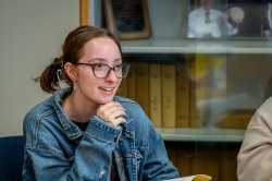 A smiling student sits in a classroom with an open book on the table in front of them.