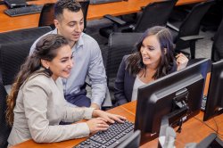 Three students in businesswear collaborate on a project in front of a multi-monitor setup