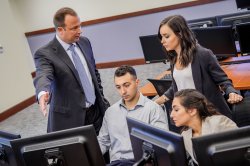 Three students and a professor all in businesswear collaborate on a project unseen behind a multiple-monitor workstation