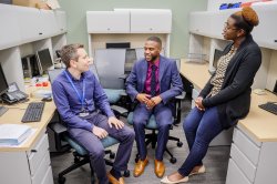 Two graduate students and one professor in professional attire have a discussion in an office space