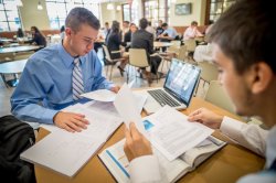 Two students in button-down shirts and ties at a cafe table with books and notes spread out and a laptop.