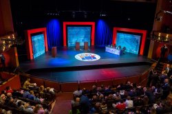 Audience members gather in a theater before a stage set for a debate with podiums, three red archways and the seal of the State of New Jersey on the stage floor.