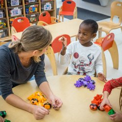 A young teacher sits at a low table with pre-school students and assembles shapes with colorful blocks with them