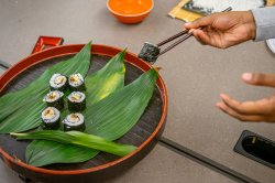 A close-up of a person using chopsticks to pick up a sushi roll from a round tray lined with large green Aspidistra leaves, with several pieces of sushi arranged on the tray. In the background, a bamboo sushi rolling mat with rice and an orange bowl are visible on the table.