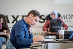 Student wearing earbuds, siting at a table and writing in notebook