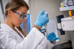 Woman student in lab coat with safety glasses and gloves uses a pipette to place a sample into a vial
