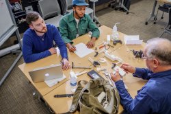 Two students and a professor sit at a work bench with a laptop and an assortment of 3D printed parts