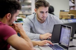 Two students sit at a table in the cafeteria intently working on their laptops