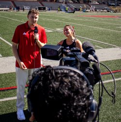 A male reporter in a red polo shirt interviews a female soccer player on the sideline at Sprague Field