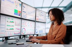 Woman in office sits at desk with six monitors displaying assorted charts and graphs