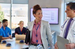 Woman in business wear with lanyard carrying a laptop under her arm chats with a doctor in a lab coat as they exit a conference room together