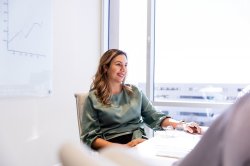 Woman in green blouse sits at desk with pen in hand speaking with a colleague off screen.
