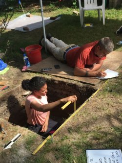 one student in an excavated hole taking measurements and another on the ground taking notes