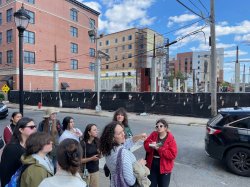 a group of students stand on the sidewalk in front of a construction site. Students take notes on walking tour with University of Orange