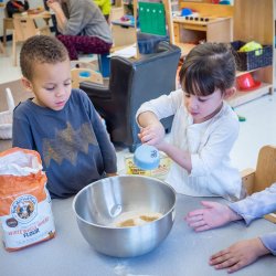 Photo of children using mixing bowls and measuring cups to make a mixture of something.