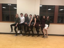 Eigth people up against a wall side by side, and they all have their left leg in the air.