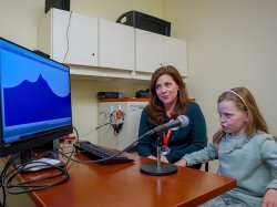 8-year-old Colette Fougere says 'r' into a microphone while Alexis Gallagher looks on.