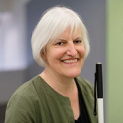 Image from the chest up of the speaker, Georgina Kleege, shows a woman with chin-length white hair from the chest up, wearing a green shirt, smiling, and carrying a long white cane.
