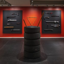 This photo features a red wall with various weapons hanging on it. There are black gym mats on the floor and a stack of 4 tires in the middle of the image with bamboo sticks in the center of the tires