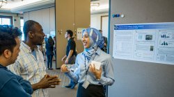 photo of students at symposium. student standing and smiling in front of research poster while talking to two attendees