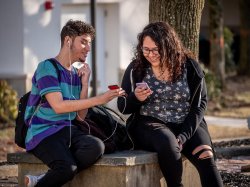 Photo of two students sitting outside playing on their phones.