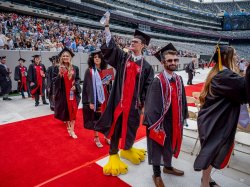 students in cap and gownstand in line as they process during Commencement ceremony. Studen Kyle Cashin stands at center, wearing large yellow, talloned feet from the University's mascot costume