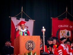 Photo of graduate Danielle McDonald in a red cap and gow. She stands behind the podium, delivering a speech as the graduate student speaker
