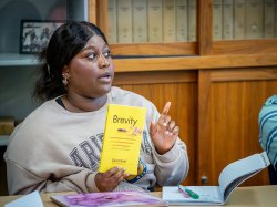 "A student gestures, as she asks a question while holding a book titled Brevity."