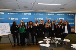 a group stands together for a photo with NYPD backdrop behind. They are all holding up the ASL sign for I love yo