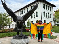 photo of Mariah Ayscue with his arms spread out revealing a yellow and rainbow colored shawl. Standing next to Red Hawk Statue, ,