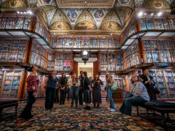 a group of students stands in the grand library room at the Morgan Library, looking in awe at the stacks of books