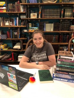 photo of student Ellie at table with laptop and stacks of books. A full bookshelf is in the background an