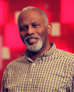 Close up photo of a man with a beard wearing a dress shirt, standing against a red backdrop