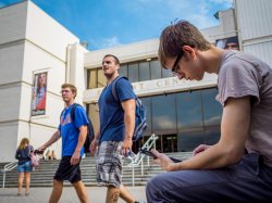Two students walking past the Student Center and a third sitting looking at his phone