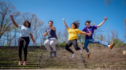 Four students wearing different t-shirts representing their College or School jumping in the University Amphitheater