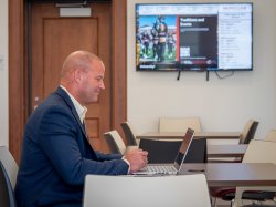 Adult student doing schoolwork on laptop in Cole Hall, Atrium with digital screen behind him promoting University events.