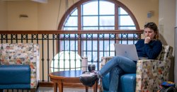 Female student sitting in chair in University Hall on laptop; window overlooking campus in the background.