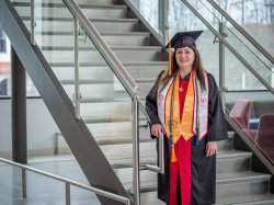 Adult degree completion student stands proudly at the foot of a staircase in Cole Hall, decorated with several graduation cords, honors and stoles in her regalia.