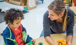 Young woman sits with a child at a low table in a kindergarten classroom