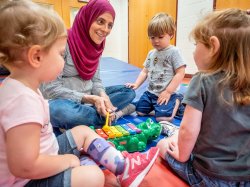 Woman sitting on mat in classroom with three toddlers showing them how to use a xylophone