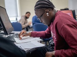 Student in maroon Bloomfield sweatshirt wears earbuds and concentrates on a collection of printed documents in front of a computer