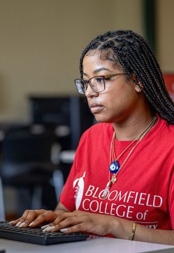 Woman student wearing red Bloomfield College shirt using computer in computer lab